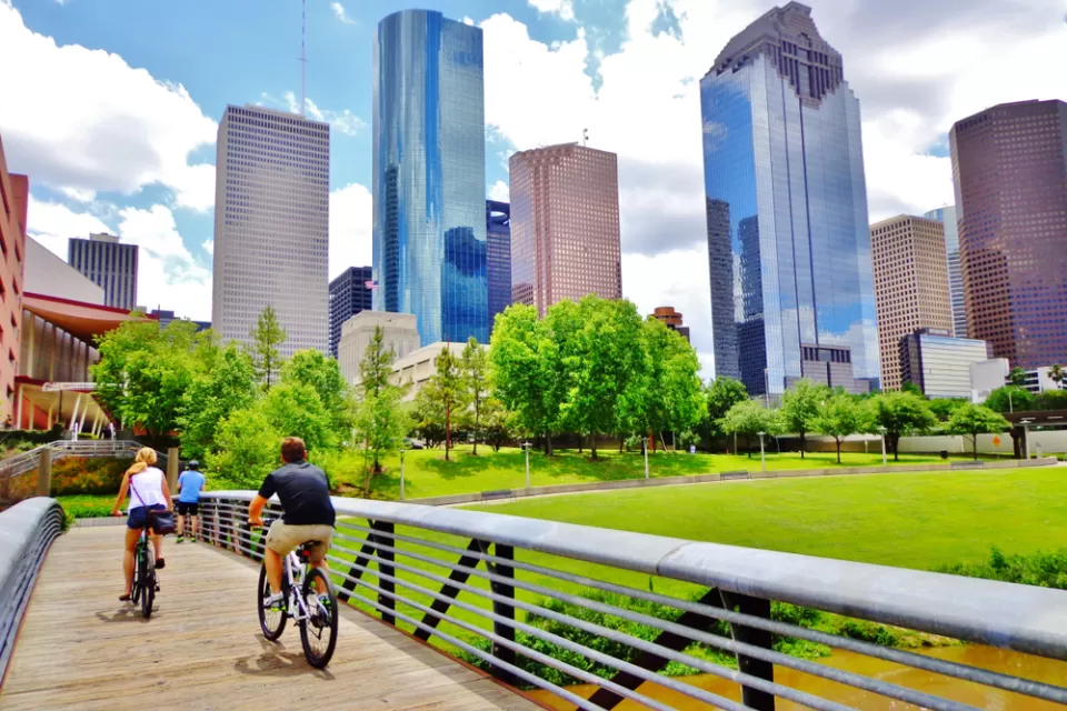 Two cyclists on a wooden bike bridge across a bayou with downtown Houston skyline in background